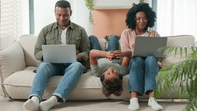 African American family Caucasian child little boy son with diverse devices multiracial parents working laptops at home happy kid lying upside down on sofa use mobile phone listen music at headphones - Powered by Shutterstock - Get 15% off with code: PIKWIZARD15