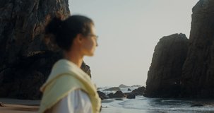A woman stands on a sandy beach at the foot of a cliff and looks into the sea. The woman's face is blurred