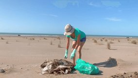 Mature woman with white hat and green gloves folding a large nylon picked up on the beach and put it in a garbage bag.