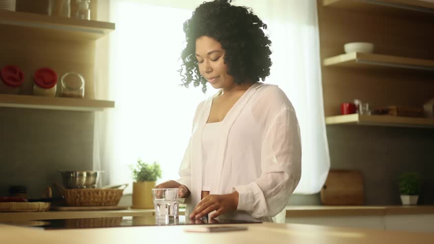 Smiling young curly woman take drug and drinking glass of still water taking medicine at home Pretty female holding pill from headache dietary supplement health care medicine for disease indoors  Royalty-Free Stock Footage #1107807147