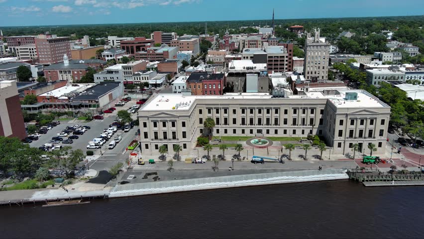 sideways aerial footage of office buildings, stores and apartments in the city skyline on Cape Fear River along the Wilmington Riverwalk, green trees, blue sky and clouds in Wilmington North Carolina