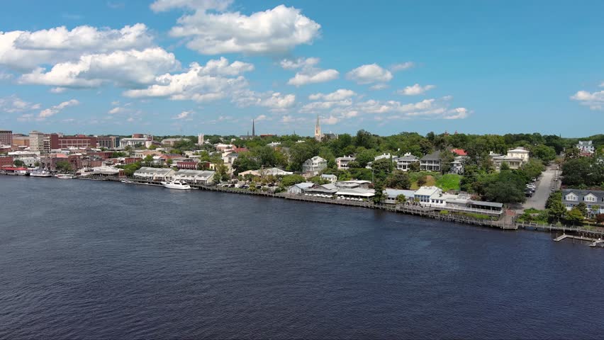  aerial footage flying along flowing waters of Cape Fear river and the Wilmington Riverwalk, office buildings and shops, lush green trees and grass with blue sky and clouds in Wilmington
