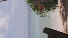 Vertical video. Little girl in straw hat and blue sundress enjoying ocean view while swinging on beach swing. Vacation and travel concept