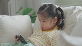 Asian child girl looking using and touch mobile phone screen on couch sofa. Baby smiling funny time to use mobile phone. Too much screen time. Cute girl watching videos while tv, Internet addiction.