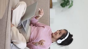 Young asian woman using laptop with earphones sitting on sofa at home.	
