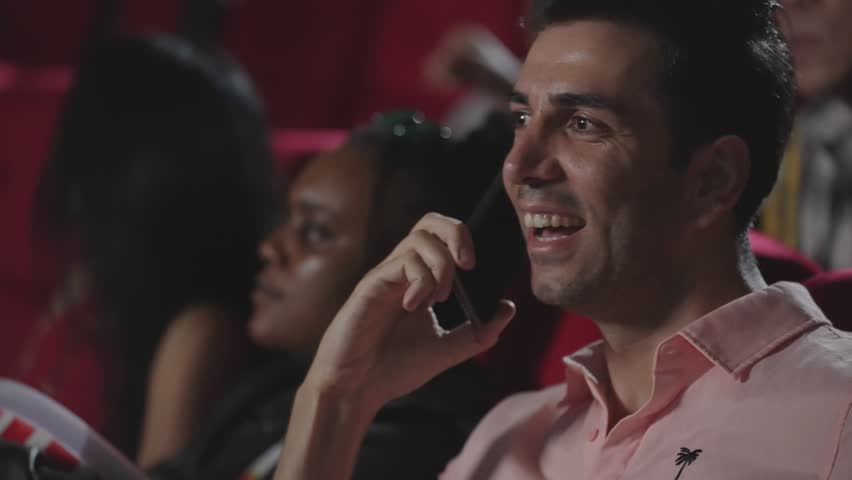 Young hispanic latin man talking on smartphone while watching movie at the cinema and disturbing audience. Phone talks prohibited during movie show. A man disrespectful talking on phone at theater