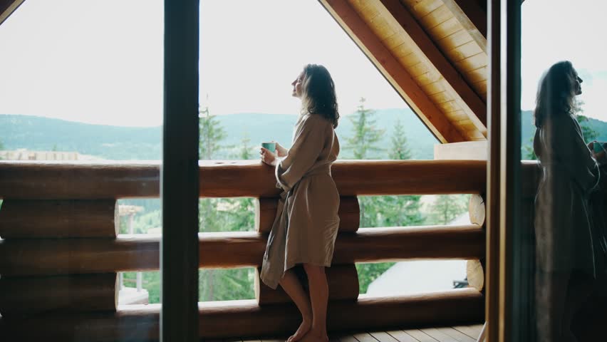 A young woman in a bathrobe stands on the terrace of a country log house and enjoys her morning coffee and fresh mountain air. Royalty-Free Stock Footage #1107840645