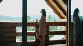 A young woman in a bathrobe stands on the terrace of a country log house and enjoys her morning coffee and fresh mountain air. - Powered by Shutterstock - Get 15% off with code: PIKWIZARD15