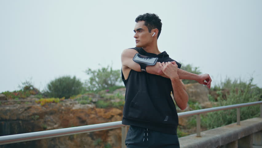 Muscular athlete warming up before race outdoors. Focused fit man stretching arms listening music in at cloudy sky. Handsome brunette sportsman training running in fresh air. Confident athletic male.