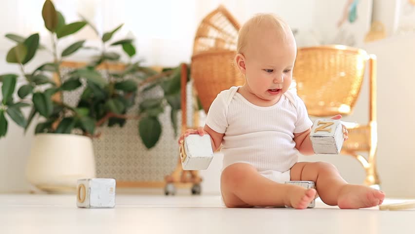 a little baby girl of six months with blue eyes in a white bodysuit plays on the floor of the children's room with wooden toys cubes smiles and rejoices, early development of the child Royalty-Free Stock Footage #1107842605