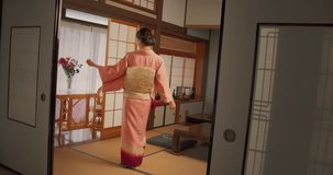 Japanese Female Gently Looking at Camera, Softly Smiling, Wearing a Traditional Pink Kimono with Flower Ornaments. Young Beautiful Woman Dancing, Posing at Home in the Living Room. Slow Motion Footage