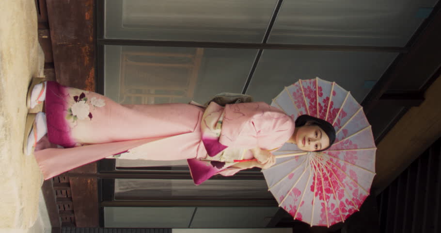 Vertical Screen: Japanese Female Looking at Camera, Softly Smiling, Wearing a Traditional Pink Kimono with Flower Ornaments and Holding an Umbrella. Young Beautiful Woman Posing Outside in Japan