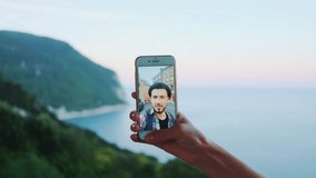 Close-up of hand holding smartphone during video call with man in front of the sea. Beautiful landscape in the background.