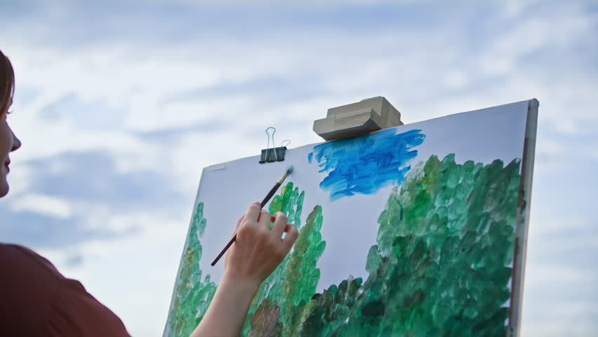 hobby, talented female artist using paints and brushes draws a picture on canvas while standing in a park against a blue sky
