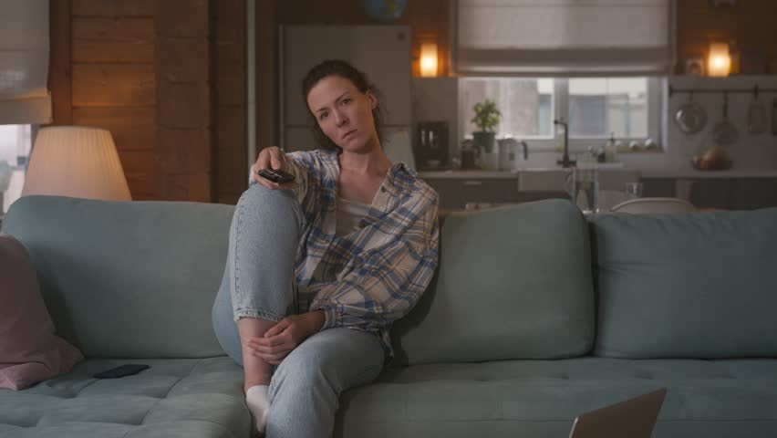 alone frustrated young woman sits on sofa at home and holds tv remote, changing channels monotonously. looks bored, uninterested and depressed.