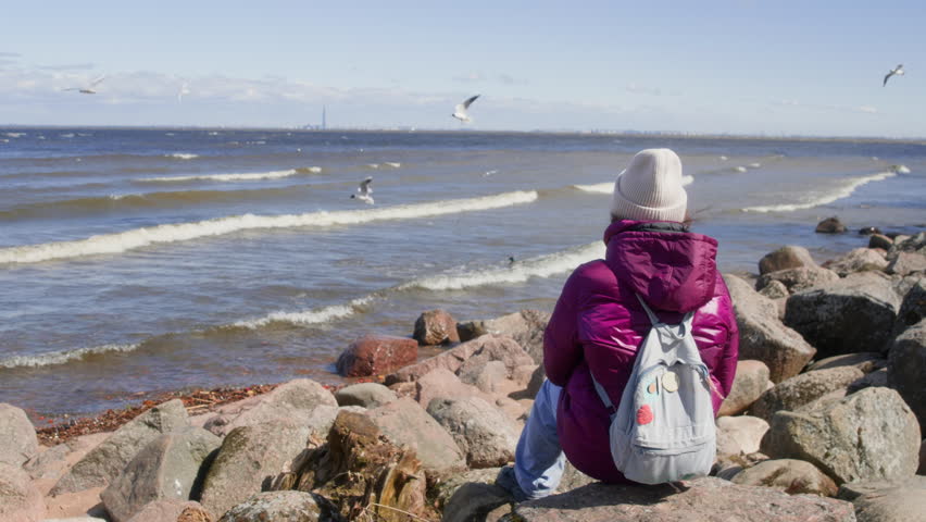Woman looking at sea waves while sitting on rock. Handheld shot of female enjoying view of beautiful seascape against sky. She is carrying backpack while spending leisure time.