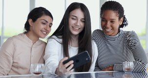 Group of happy beautiful multiethnic girls making video call on mobile phone, using cellphone at table in coffee shop, drinking beverages, watching online content, enjoying Internet communication