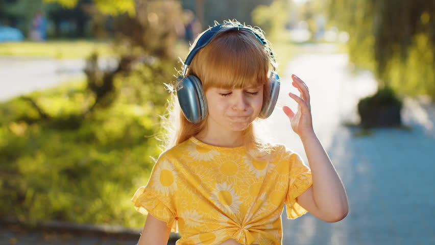 Happy smiling preteen child kid taking off wireless headphones choosing, listening energetic disco music in smartphone dancing. Blonde funny girl looking at camera standing on city sunset park street Royalty-Free Stock Footage #1107893603