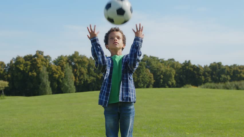 Portrait of positive cute school age African American boy in casual clothes posing , playing and tossing up soccer ball up in the air, looking with cheerful radiant smile on green field. Royalty-Free Stock Footage #1107897667