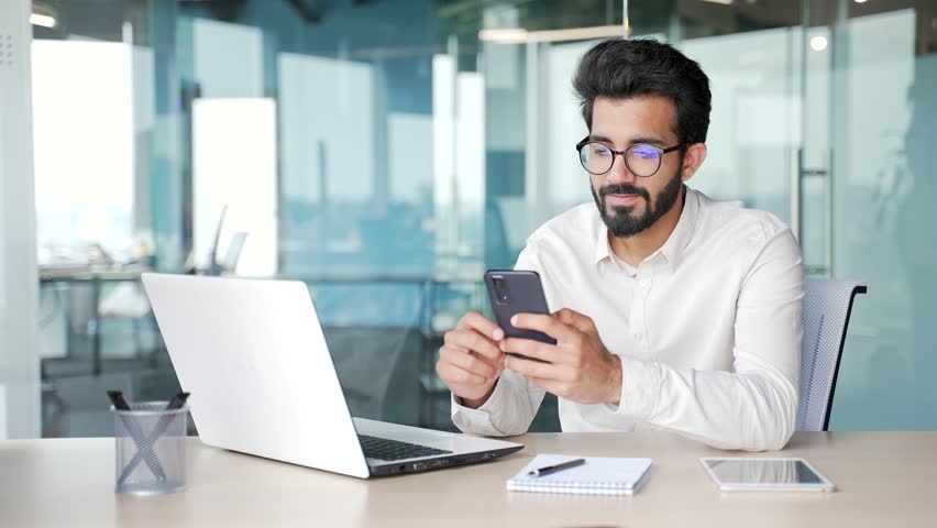 A smiling young businessman in a white shirt is using, browsing smartphone while sitting at workplace in modern office. Bearded entrepreneur in glasses checking email or writing text message to client | Shutterstock HD Video #1107898585