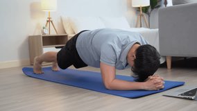 happy asian young man doing exercise and watching tutorial on laptop with mat in living room at home. Asia male doing plank workout