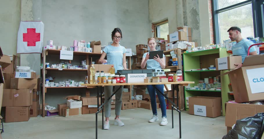 Volunteer with digital tablet in hands standing near young couple packing cardboard boxes with foodstuff at warehouse. Team of volunteers preparing donation for red cross organization.