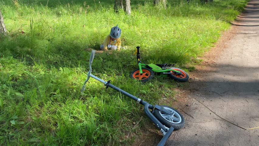 child in safety helmet with balance bike and toy camera on walk in summer park explores a mole hole