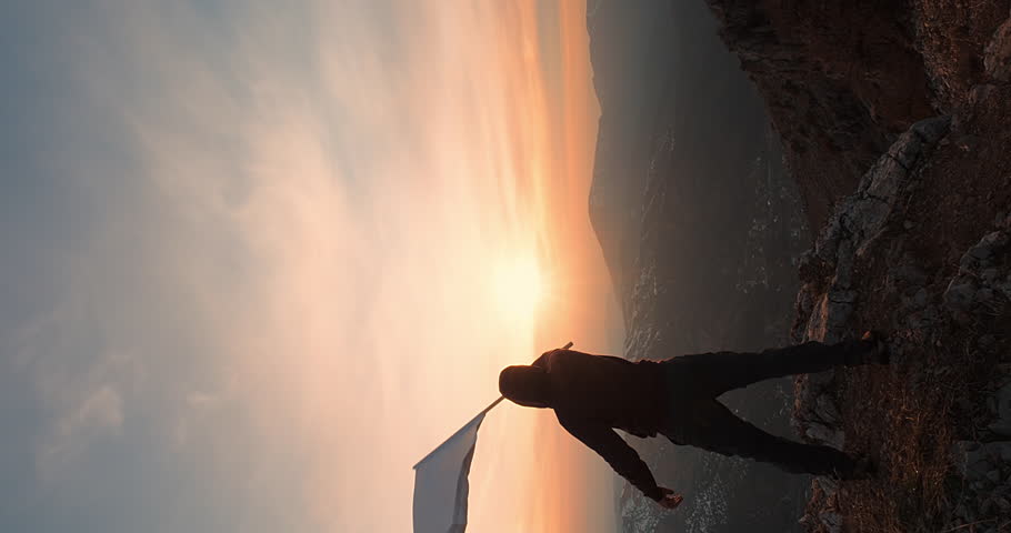 Vertical shot of a Strong Man on Top of a Mountain with Cinematic Sunset Background Waving a White Flag while the Wind Blows. Concept of World Peace, Say No to War, Conquering Goals and Dreams.. Royalty-Free Stock Footage #1107930623