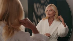 Middle-aged Caucasian old mature woman in bathrobe looking at mirror combing clean hair with hairbrush in home bathroom beautiful senior lady self care beauty routine natural shampoo balm conditioner - Powered by Shutterstock - Get 15% off with code: PIKWIZARD15