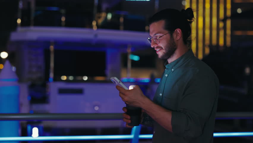Side View of the Handsome Happy Guy Using Smartphone While Walking on Night City Street in Casual Clothes. Smiling Man Typing, Browsing, and Using Mobile Phone in His Hands. Outside, Outdoors Royalty-Free Stock Footage #1107931637