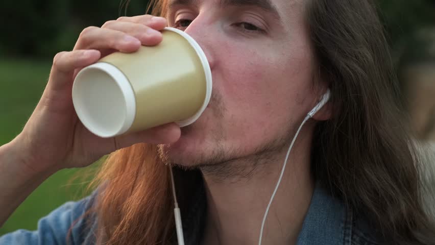 Young man with headphones smoking e-cigarette and drinking coffee outdoor at evening | Shutterstock HD Video #1107931915