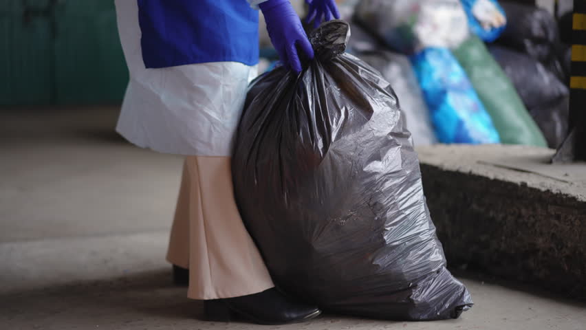 Unrecognizable female volunteer placing garbage bags sorting trash on recycling station. Young Caucasian woman in uniform walking with rubbish indoors