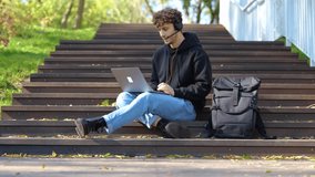 Happy guy working online with laptop while sitting on the staircase, using headset in the park. Autumn season. Education, working concept. Slow motion