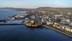 Medieval Norman Castle and harbor in Carrickfergus near Belfast, Northern Ireland, in sunrise light. Aerial 4K approaching video with marina, yachts, breakwater and town