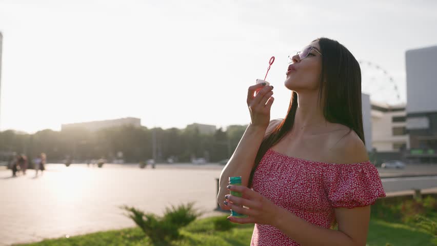 Beautiful Woman in Glasses Blowing Soap Bubbles Outdoors. Slow Motion Shot of Childhood Time in Adult Age. Fun Under the Rays of the Sun. Portrait of Happy Girl Playing Childish Game. 