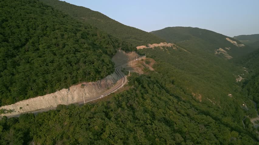 Drone video captures a car driving through an uphill mountain pass during sunset. The road is framed by lush green forest, creating a serene and scenic journey. Perfect for travel, adventure, and natu