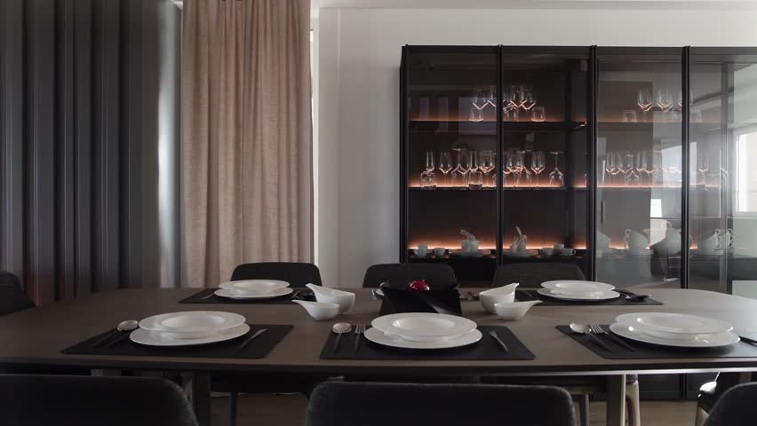 Stylish table setting in dining room at modern apartment with minimalist design. Empty clean white plates and silver cutlery on black placemats on wooden tabletop. Dinner party concept | Shutterstock HD Video #1107944225