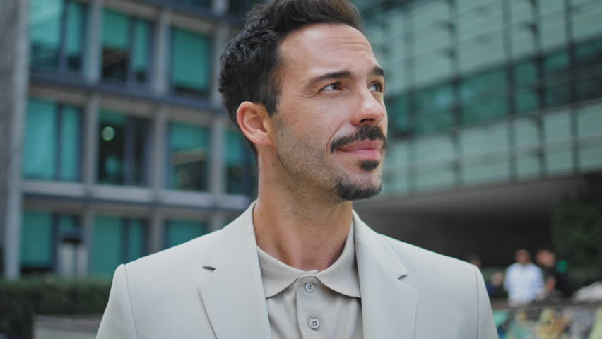 Smiling ceo posing downtown portrait. Young brunette man looking camera standing in glass architecture background close up. Relaxed hispanic guy staring distance waiting alone at street zoom out