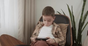 Cute school age boy using digital phone device, kid sitting alone on sofa with gadget. The concept of children's technical addiction. Internet and playing the game at home. 4K.
