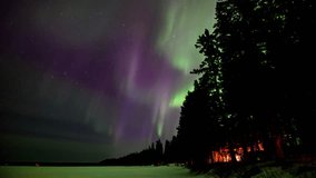 View of bright green and purple Aurora in a star filled sky. The foreground includes a snow-covered lake and a shoreline of silhouettes of pine and spruce trees. Orange colored lights come from cabins - Powered by Shutterstock - Get 15% off with code: PIKWIZARD15