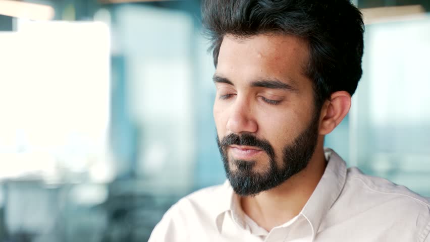 Close up A young bearded man in a shirt drinks water from a glass while sitting at a workplace in the office. Happy smiling employee feeling relieved, enjoying a clean cool drink, relaxing and resting Royalty-Free Stock Footage #1107954689