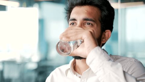 Close up A young bearded man in a shirt drinks water from a glass while sitting at a workplace in the office. Happy smiling employee feeling relieved, enjoying a clean cool drink, relaxing and resting Arkivvideo