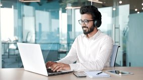 Young smiling IT specialist coder in wireless headphones typing on laptop computer while sitting at workplace at desk in modern office. A handsome smiling developer in a white shirt works on a project - Powered by Shutterstock - Get 15% off with code: PIKWIZARD15