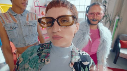 Short-haired girl with nose piercing and tattoos wearing trendy outfit posing like a model on camera with queer dance crew in the background Stockvideó