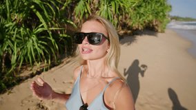 Girl video blogger filming herself on selfie camera with view of ocean, sandy beach and palm trees. Woman traveler in sunglasses walking at shore recording video about adventure on tropical island.