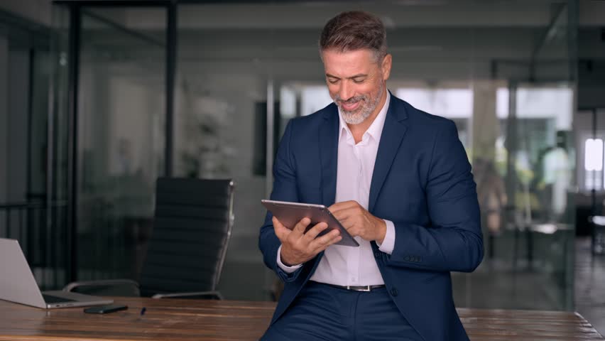 Happy Latin Hispanic bearded stylish mature adult professional business man, smiling Indian senior businessman CEO holding digital tablet using fintech tab application standing inside company office. Royalty-Free Stock Footage #1107956615