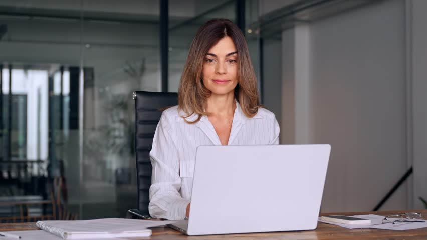 Smiling European Latin business woman accountant analyst holding documents, work at laptop computer doing online trade market tech research. Focused Hispanic businesswoman with paperwork in office. Royalty-Free Stock Footage #1107956625