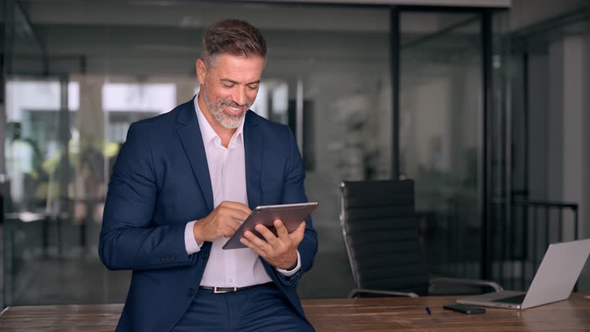 Happy Latin Hispanic bearded stylish mature adult professional business man, smiling Indian senior businessman CEO holding digital tablet using fintech tab application standing inside company office. Royalty-Free Stock Footage #1107956631