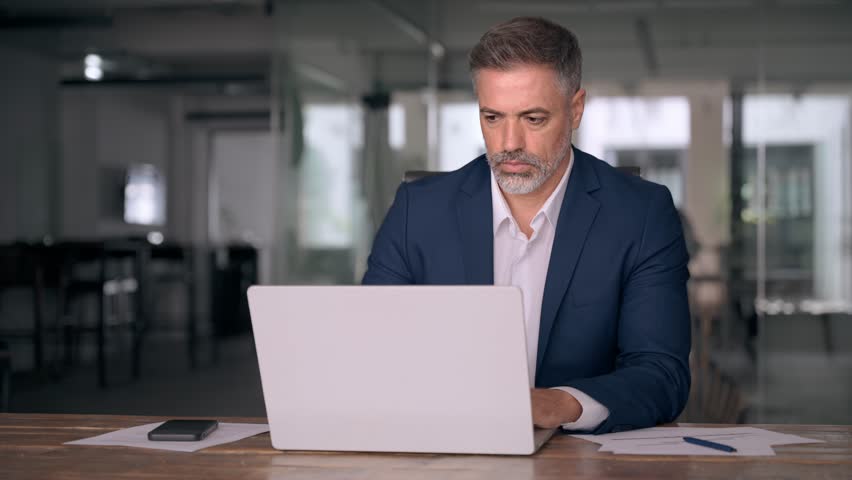 Mature Indian or Latin business man ceo trader using computer, typing, working in modern office, doing online data market analysis, thinking planning tech strategy looking at laptop with copy space. Royalty-Free Stock Footage #1107956635