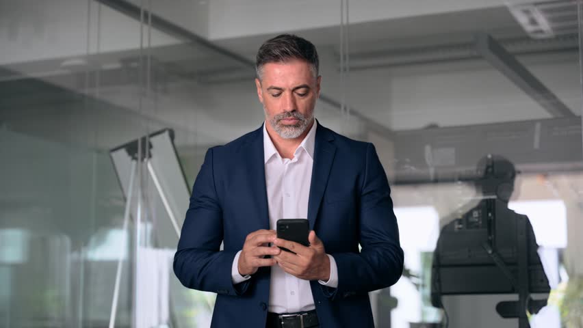Mature Latin or Indian businessman walking with smartphone in office. Middle aged manager using cell phone mobile app. Digital technology application and solutions for business success development. Royalty-Free Stock Footage #1107956637